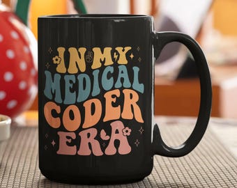 May include: A black ceramic mug featuring the text "IN MY MEDICAL CODER ERA" in a multi-colored, wavy font. The design includes small flower and star icons interspersed among the words, with the lettering in shades of yellow, blue, orange, and pink.