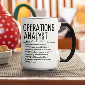 Op de afbeelding: Een witte keramische koffiemok met een zwarte rand en handvat. De mok heeft een zwart-witte afbeelding met de tekst "OPERATIONS ANALYST noun(n.) Jessica The wizards of efficiency, conjuring up algorithms and crunching numbers to optimise everything from supply chains to scheduling, all while quietly judging inefficiencies like a disappointed parent watching a messy room."