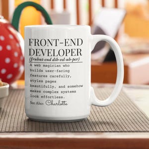 May include: A white ceramic mug featuring black printed text that defines a front-end developer as a web magician who builds user-facing features, styles pages, and makes complex systems look effortless. The text includes the name Charlotte.