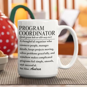 Könnte beinhalten: Ein weißer Keramikbecher mit schwarzem Aufdruck, der einen Programmkoordinator als aufmerksamen Organisator definiert, der Menschen verbindet, Details verwaltet und Probleme löst, endend mit dem Satz See Also: Andrea.