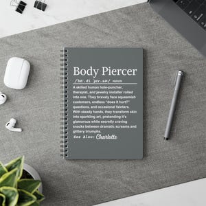 May include: A gray spiral-bound notebook with the text "Body Piercer" in white. The description below defines the term, including the phrase "See Also: Charlotte". A pen, earbuds, and other office supplies are visible.