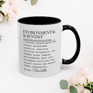 Könnte beinhalten: Weißer Keramikbecher mit schwarzem Innenraum und Henkel. Der Becher trägt den Text "ENVIRONMENTAL SCIENTIST" mit einer Definition, einschließlich des Namens "Charlotte" unten. Der Becher ist von Blumen umgeben.