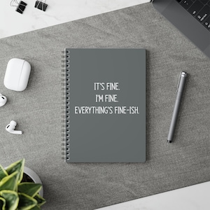 May include: A dark gray spiral-bound notebook featuring the white printed text IT'S FINE. I'M FINE. EVERYTHING'S FINE-ISH. centered on the front cover. The notebook is positioned on a flat surface alongside a gray pen, white earbuds, and a case.