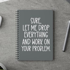 May include: A grey spiral-bound notebook featuring the white text: "Sure, Let Me Drop Everything And Work On Your Problem". The notebook is on a grey surface, with a pen and other office supplies visible.
