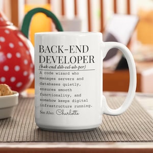 May include: White ceramic mug with the text "BACK-END DEVELOPER" and a definition. The mug also includes the name "Charlotte". The mug has a large handle and is sitting on a patterned surface.