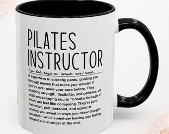 May include: A white ceramic mug with a black interior and handle, featuring black printed text that defines a Pilates instructor as a superhero in stretchy pants who guides you through moves, combining strength, flexibility, and patience.