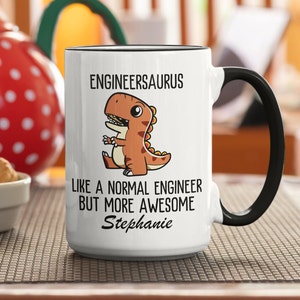May include: White ceramic coffee mug with a black rim and handle. The mug features a cartoon dinosaur with the text "Engineersaurus" above it and "Like a normal engineer but more awesome Stephanie" below it.