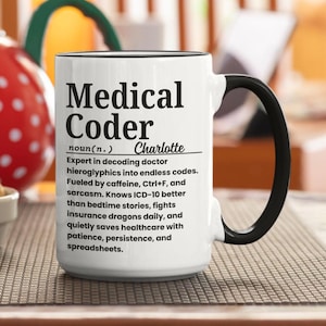 Pode incluir: Caneca de cerâmica branca com alça e borda pretas. A caneca apresenta o texto "Medical Coder" em letras pretas em negrito, juntamente com uma definição e um nome. A caneca é projetada para um codificador médico.
