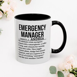May include: White ceramic mug with a black interior and handle. The mug features the text "EMERGENCY MANAGER" in bold black letters, along with a definition and a personalized name. The mug is surrounded by flowers.