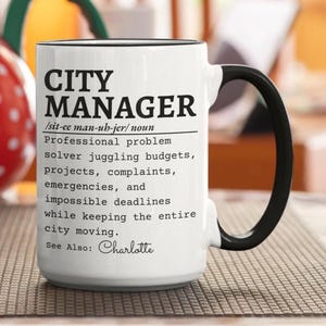 Puede incluir: Una taza de cerámica blanca con asa y borde negros, que presenta el texto impreso en negro CITY MANAGER seguido de una pronunciación fonética, una definición del rol como solucionador de problemas y el texto See Also: Charlotte.