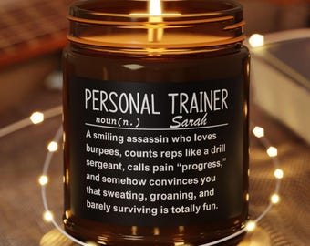 May include: A brown glass candle jar with a lit wick, resting on a wooden coaster. The jar has a black label with white text that reads "PERSONAL TRAINER" and a humorous definition. Fairy lights surround the candle.