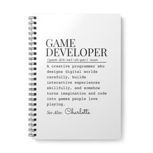 Puede incluir: Cuaderno blanco con espiral con el texto "GAME DEVELOPER" y su definición. La definición describe a un programador creativo que diseña mundos digitales. También se incluye el texto "See Also: Charlotte".
