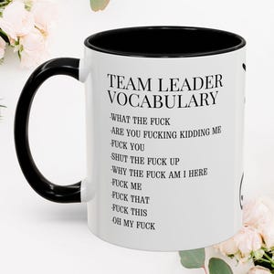 Op de afbeelding: Een witte keramische mok met een zwarte handgreep en binnenkant, voorzien van de zwarte tekst TEAM LEADER VOCABULARY gevolgd door een lijst met zinnen waaronder WHAT THE FUCK, ARE YOU FUCKING KIDDING ME, FUCK YOU, SHUT THE FUCK UP, WHY THE FUCK AM I HERE.