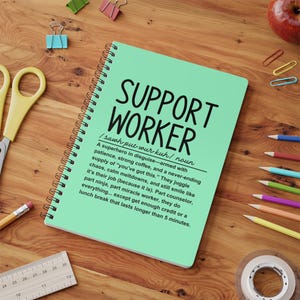 Pode incluir: Um caderno verde com espiral e as palavras "SUPPORT WORKER" em texto preto. A definição da palavra "SUPPORT WORKER" está escrita em texto preto abaixo do título. A definição diz: "Um super-herói disfarçado - armado com paciência, café forte e um suprimento infinito de "você consegue". Eles fazem malabarismos com o caos, acalmam os colapsos e ainda assim sorriem como se fosse seu trabalho (porque é). Em parte conselheiro, em parte ninja, em parte trabalhador milagroso, eles fazem tudo... exceto obter crédito suficiente ou um intervalo para o almoço que dure mais de 5 minutos."