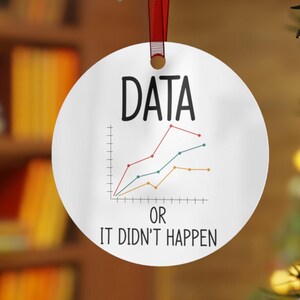 Puede incluir: Adorno navideño redondo blanco con la palabra "DATA" en negro, un gráfico de líneas y el texto "OR IT DIDN'T HAPPEN". El adorno cuelga de una cinta roja. El fondo está borroso.