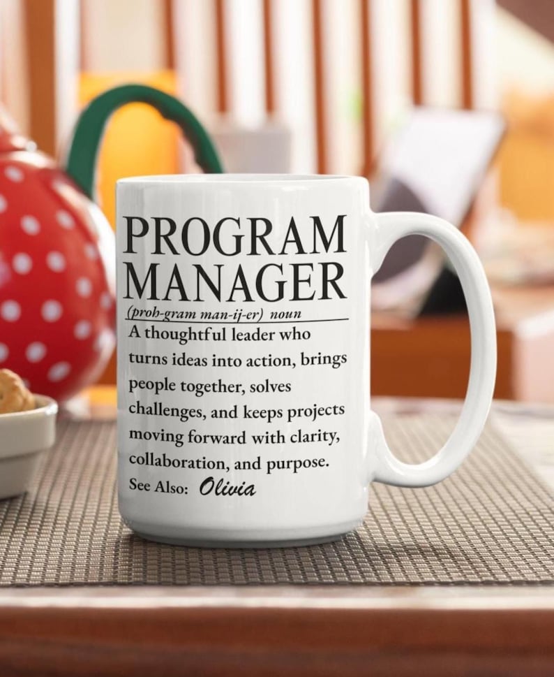 May include: A white ceramic mug featuring black printed text that defines the term Program Manager as a thoughtful leader who turns ideas into action, brings people together, solves challenges, and keeps projects moving forward. It includes the text See Also: Olivia.