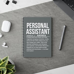 Op de afbeelding: Een grijze spiraalgebonden notitieblok met de woorden "PERSONAL ASSISTANT" in grote witte letters. De tekst hieronder definieert een persoonlijke assistent. Een pen, oortelefoons en paperclips zijn ook zichtbaar.