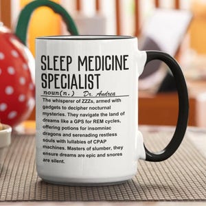 Peut inclure: Un mug en céramique blanche avec une anse et un bord noirs, portant un texte imprimé en noir : SLEEP MEDICINE SPECIALIST noun(n.) Dr. Andrea. Le texte inclut une définition humoristique décrivant un professionnel aidant les autres à dormir.