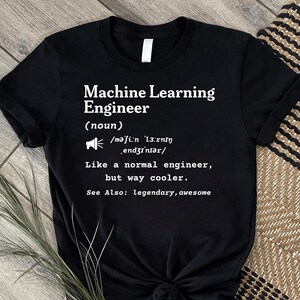 May include: A black short-sleeved t-shirt featuring white text that reads: Machine Learning Engineer (noun) /məˈʃiːn ˈlɜːrnɪŋ ˌendʒɪˈnɪər/ Like a normal engineer, but way cooler. See Also: legendary, awesome. The shirt has a crew neckline.