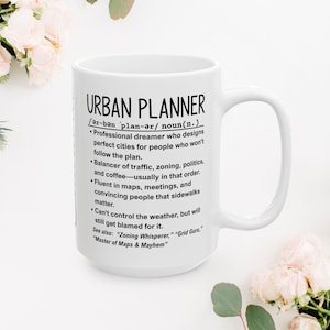 May include: A white ceramic mug featuring the text "URBAN PLANNER" and a definition. The definition includes phrases like "Professional dreamer" and "Balancer of traffic, zoning, politics, and coffee." The mug has a large handle and is surrounded by flowers.
