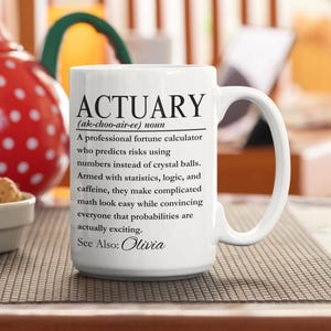 Könnte beinhalten: Ein weißer Keramikbecher mit schwarzem Aufdruck, der das Wort ACTUARY als professionellen Glücksrechner definiert, der Risiken mit Zahlen statt mit Kristallkugeln vorhersagt, bewaffnet mit Statistik, Logik und Koffein. Er enthält den Text See Also: Olivia.