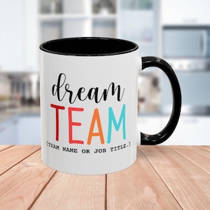 Pode incluir: Caneca de cerâmica branca com interior e alça pretos. A caneca apresenta as palavras "dream TEAM" numa mistura de cores, com o texto "(TEAM NAME OR JOB TITLE.)" por baixo.