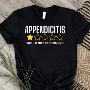 Puede incluir: Camiseta negra con la palabra "APENDICITIS" en blanco, con una estrella amarilla y cuatro estrellas delineadas debajo. El texto "NO RECOMENDARÍA" está debajo de las estrellas. La camiseta está sobre una superficie de madera.