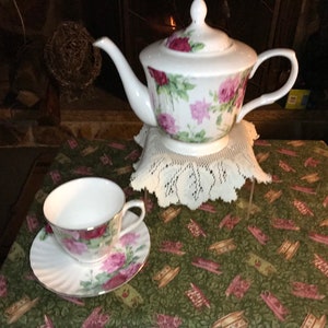 May include: A white teapot and teacup set with a floral pattern featuring pink roses and green leaves. The teapot has a lid and a handle. The teacup has a saucer.