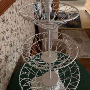 Könnte beinhalten: Weißer dreistöckiger Drahtständer mit einem dekorativen Abschluss oben. Jede Stufe hat einen runden Drahtkorb und eine weiße Holzbasis.