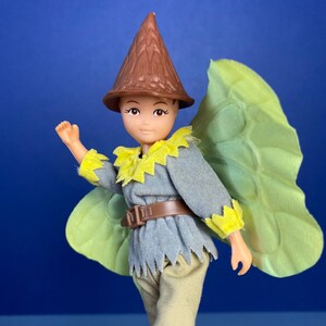 May include: A plastic doll wearing a brown pointed hat, a grey shirt with yellow trim, a brown belt, and beige trousers. The doll has a green fabric butterfly wing attached to its back.