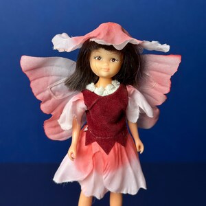 May include: A pink fairy doll with brown hair, wearing a pink flower hat and a burgundy dress with pink flower wings.