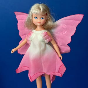 May include: A vintage doll with blonde hair and blue eyes, dressed in a white and pink dress, and pink butterfly wings. The doll has a sweet expression against a solid blue background.