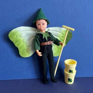 May include: A vintage fairy doll wearing a green pointed hat, green and black outfit, and large leaf-like wings. The doll holds a small rake and stands next to two yellow flower pots decorated with blue floral designs. The doll has brown hair and a brown belt.