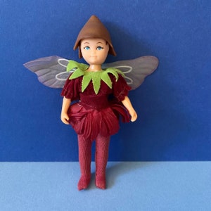 Peut inclure: Une petite poupée habillée en fée avec un chapeau brun, un col vert et une jupe rouge en forme de fleur. La fée a les yeux bleus et un visage rose clair. La poupée a des ailes en matériau violet translucide.