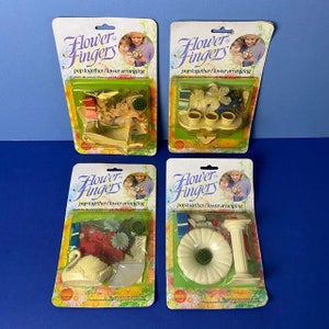 May include: Four packs of Flower Fingers pop-together flower arranging kits. Each pack features a different flower arrangement, including a swan, a daisy, a flower pot, and a vase. The packs are all yellow and green with the Flower Fingers logo.