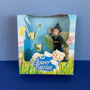 May include: A green and brown Flower Fairies doll with a green hat and wings. The doll is holding a rake and a bucket. The doll is packaged in a blue and white box with the Flower Fairies logo.