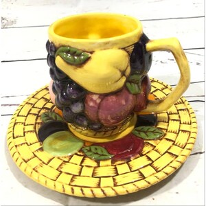 May include: A yellow ceramic teacup and saucer set with a fruit design. The cup features a pear, grapes, and a peach. The saucer has a woven basket design with fruit accents.