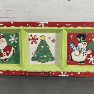 May include: A ceramic serving tray with three divided sections, each featuring a different Christmas scene. The left section has Santa Claus carrying presents, the middle section has a Christmas tree, and the right section has a snowman holding a small Christmas tree. The tray has a red and green border with white snowflakes.