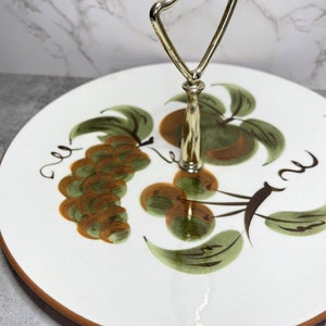 May include: A white ceramic serving tray with a gold handle. The tray features a hand-painted design of green leaves and orange fruit.