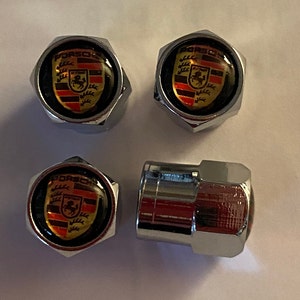 May include: Four chrome-finished tyre valve stem caps. Each cap displays a Porsche crest logo in black, red, and gold. The caps are hexagonal and have a shiny, reflective surface.