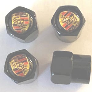 May include: Four black Porsche wheel nuts. Each nut features the Porsche logo, a gold shield with a black horse and red and black stripes. The nuts are hexagonal.