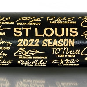 May include: A black baseball bat with gold lettering. The bat is signed by the St. Louis Cardinals players from the 2022 season. The text on the bat reads "Players Officially Licensed Product of MLB Players Inc. Limited Edition 102 of 5000".