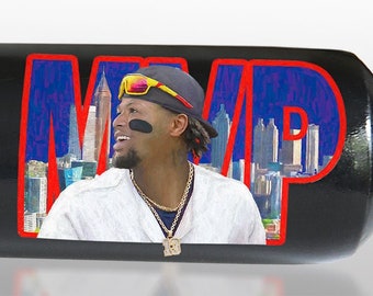 May include: A black baseball bat with gold lettering. The bat is a limited edition, number 23 of 2023. The bat features a photo of baseball player Ronald Acuña Jr. with the text "2023 Most Valuable Player" and his stats. The bat is a Players product.