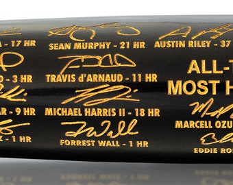 May include: A black baseball bat with gold lettering commemorating the Atlanta Braves' 2023 all-time league record for most home runs in a season, with a total of 307 home runs. The bat features signatures from players who contributed to the record, including Orlando Arcia, Sean Murphy, Austin Riley, Matt Olson, Sam Hilliard, Travis d'Arnaud, Kevin Pillar, Michael Harris II, Marcell Ozuna, Ronald Acuña Jr., and Forrest Wall.