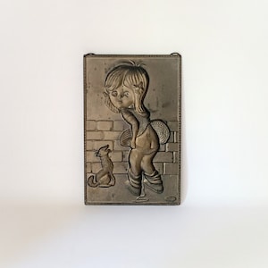Może przedstawiać: Metalowa tabliczka ścienna przedstawiająca dziewczynkę i kota. Dziewczynka ma na sobie sukienkę i ma włosy w warkoczykach. Kot siedzi na ziemi i patrzy na dziewczynkę. Tabliczka jest prostokątna i ma brązowe wykończenie.