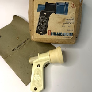 May include: An off-white vintage pelmeni maker with a handle and nozzle, next to its original packaging. The packaging features a diagram of the tool and Cyrillic text. A matching envelope is also present.