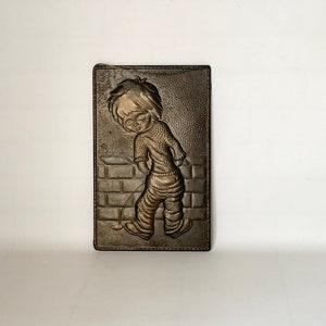 May include: A bronze wall plaque with a raised image of a child peeing on a brick wall. The child is wearing a sweater and pants.
