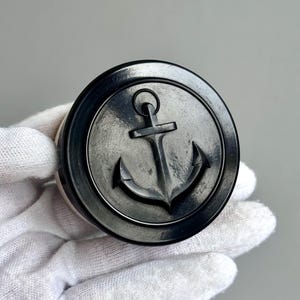 Puede incluir: Un tapón de botella redondo, gris oscuro, con un diseño de ancla en relieve en el centro. El ancla es un símbolo náutico clásico, y el tapón tiene un brillo metálico. El tapón se sostiene en una mano enguantada.