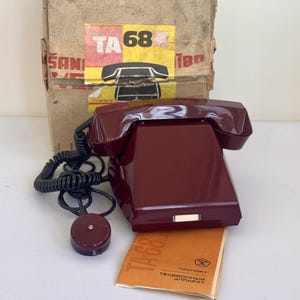 May include: A vintage burgundy telephone with a coiled cord and a separate earpiece. The phone rests on a small orange booklet. A cardboard box with a yellow and red label is in the background. The label reads "TA 68".