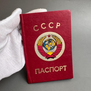 May include: A red Soviet passport featuring the gold embossed text CCCP at the top and ПАСПОРТ at the bottom. In the center is a circular emblem displaying a globe, a hammer and sickle, wheat stalks, and a red star on a white background.